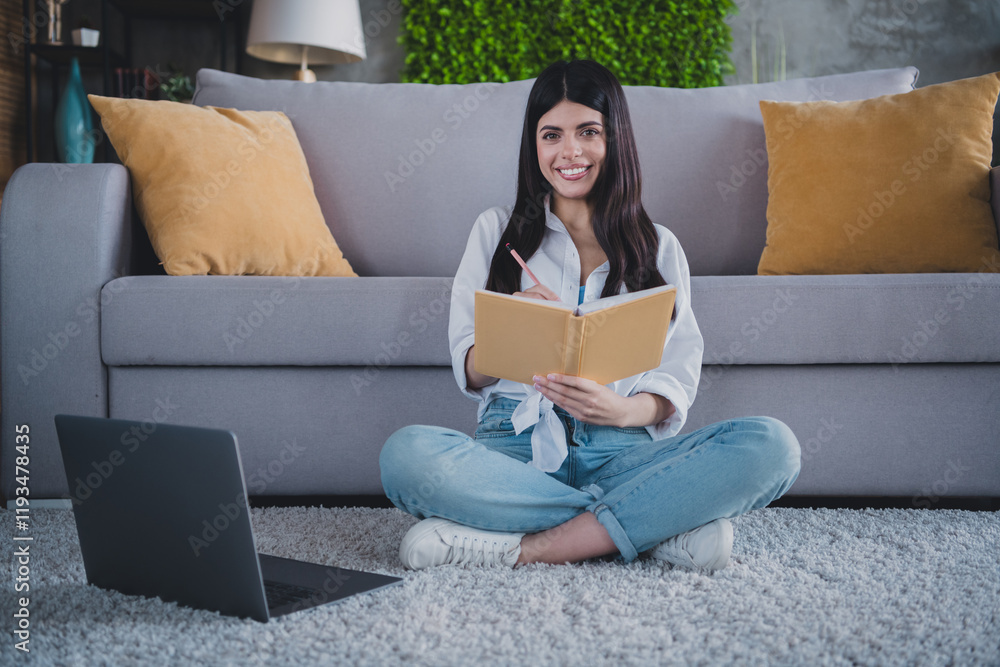 Young woman sitting on floor indoors writing in a notebook with a laptop nearby Young woman sitting on floor indoors writing in a notebook with a laptop nearby