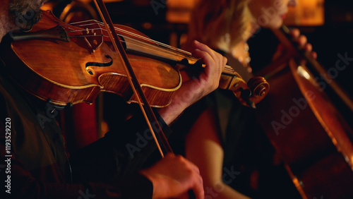 A close-up of musicians playing string instruments in an orchestra, focusing on a violinist and a cellist. The warm lighting highlights the wood of the instruments.