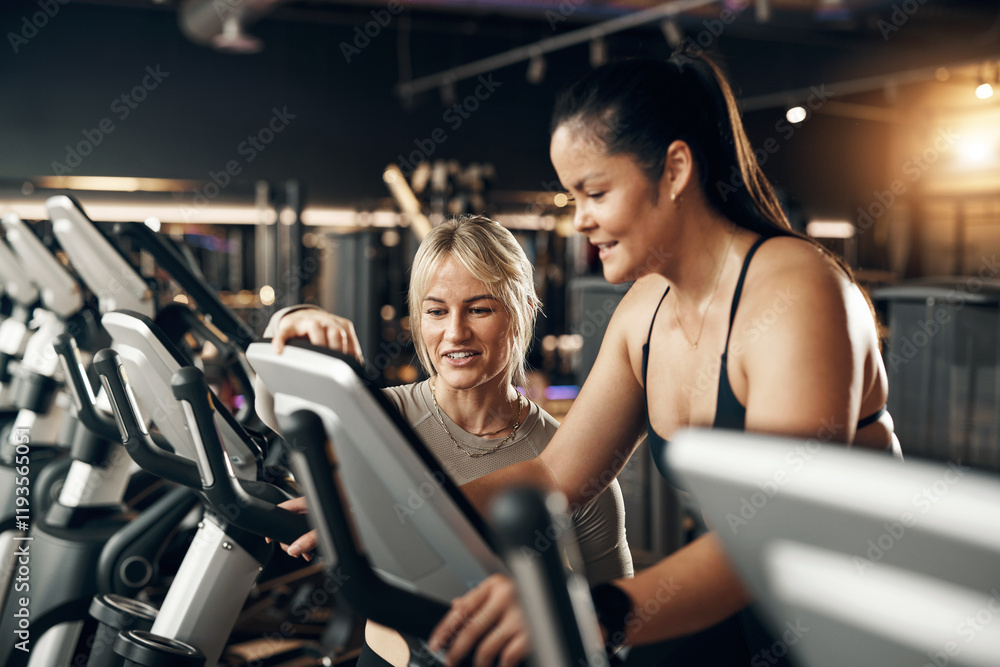 Smiling female instructor offering encouragement to a fit young woman riding on a stationary bike during a cardio exercise session at the gym Smiling female instructor offering encouragement to a fit young woman riding on a stationary bike during a cardio exercise session at the gym