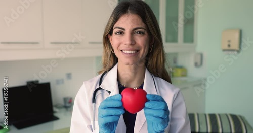 Beautiful doctor woman promoting heart checkup, cardiology examination, donation. Happy young Hispanic cardiologist showing toy red heart, looking at camera, smiling. Professional portrait photo