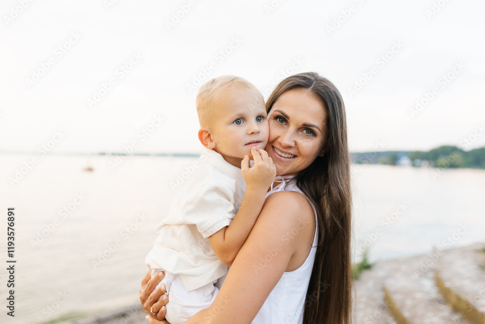 Happy woman is holding baby in her arms while standing on beach. Concept of warmth and happiness Happy woman is holding baby in her arms while standing on beach. Concept of warmth and happiness