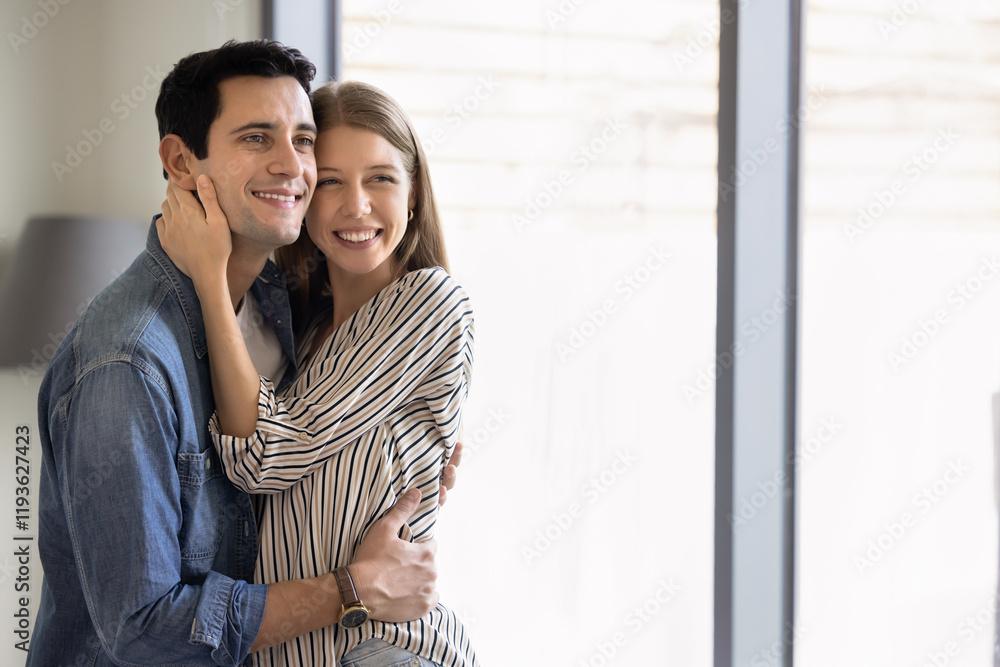 Happy attractive young married couple hugging at home with window and daylight in background, looking at copy space away, smiling, posing for casual portrait Happy attractive young married couple hugging at home with window and daylight in background, looking at copy space away, smiling, posing for casual portrait
