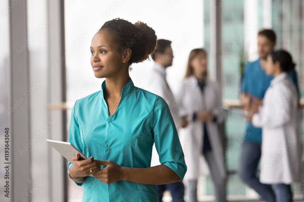 Attractive African female nurse or medical assistant standing in clinic hallway with digital tablet device looks out window with thoughtful expression, deep in reflection, decision-making. Healthcare Attractive African female nurse or medical assistant standing in clinic hallway with digital tablet device looks out window with thoughtful expression, deep in reflection, decision-making. Healthcare