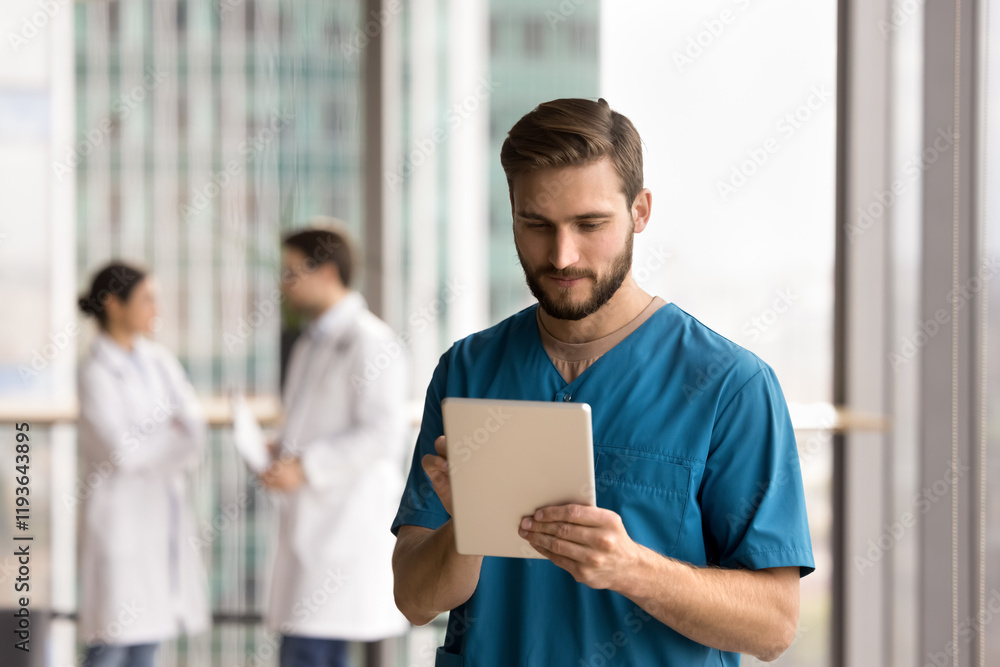 Focused man medical worker in blue uniform using modern digital tablet at workplace, reviewing electronic health records, scheduling patient appointments via application, messaging, coordinating staff Focused man medical worker in blue uniform using modern digital tablet at workplace, reviewing electronic health records, scheduling patient appointments via application, messaging, coordinating staff