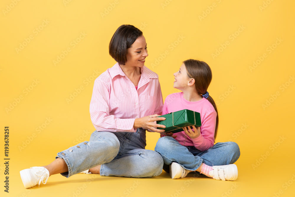 A mother sits on the floor with her daughter, smiling and exchanging a green gift box. They are surrounded by a bright yellow background, showcasing their joyful bond and affection. A mother sits on the floor with her daughter, smiling and exchanging a green gift box. They are surrounded by a bright yellow background, showcasing their joyful bond and affection.