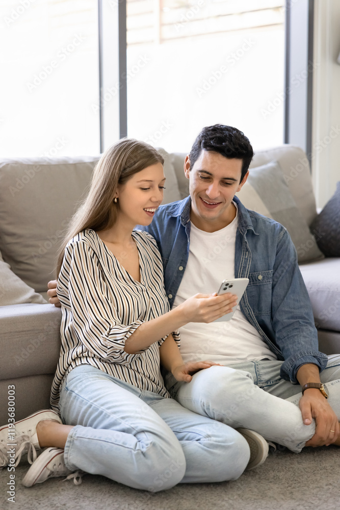 Happy young Hispanic couple resting on heating floor at home, using smart application on mobile phone, enjoying modern technology, online communication, browsing booking website, shopping Happy young Hispanic couple resting on heating floor at home, using smart application on mobile phone, enjoying modern technology, online communication, browsing booking website, shopping