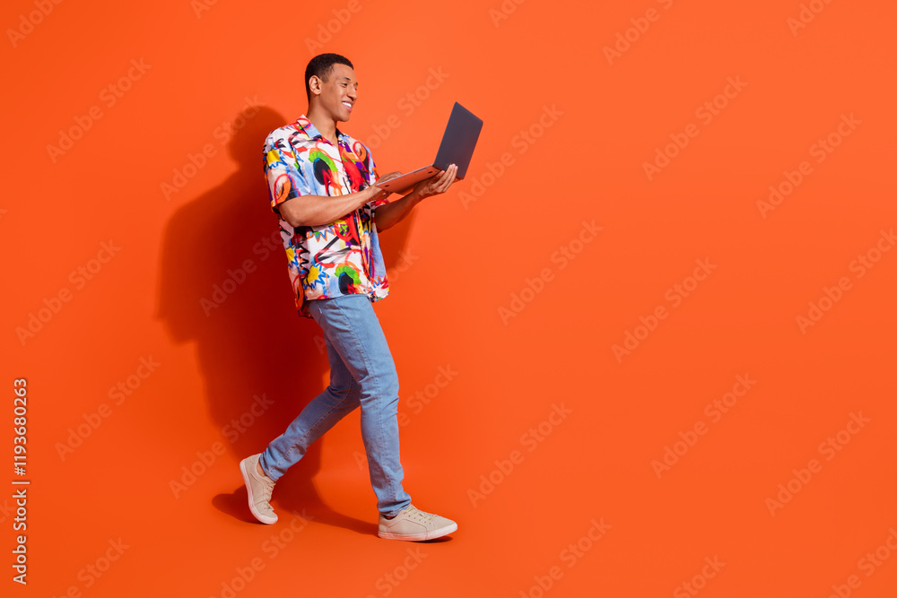 A confident young man in a vibrant, stylish shirt walks against an orange background while looking at his laptop. A confident young man in a vibrant, stylish shirt walks against an orange background while looking at his laptop.