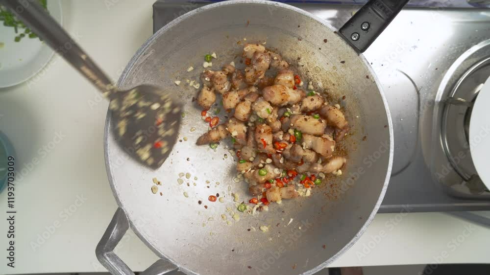 Chef adds seasoning from a small white bowl to a wok containing pork belly stir-fry, garlic, chili peppers, and other spices, creating a flavorful and aromatic dish