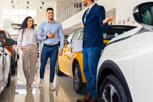 Middle-eastern luxury car salon concept. Cheerful arab man sales assistant giding young family husband and wife by huge showroom with luxury automobiles, salesman showing appropriate auto, full length photo