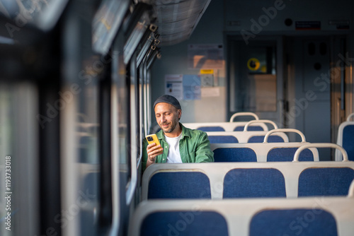 Bilde på lerret Smiling man using smartphone, scrolling social media and messaging, seated by the window in train in the morning