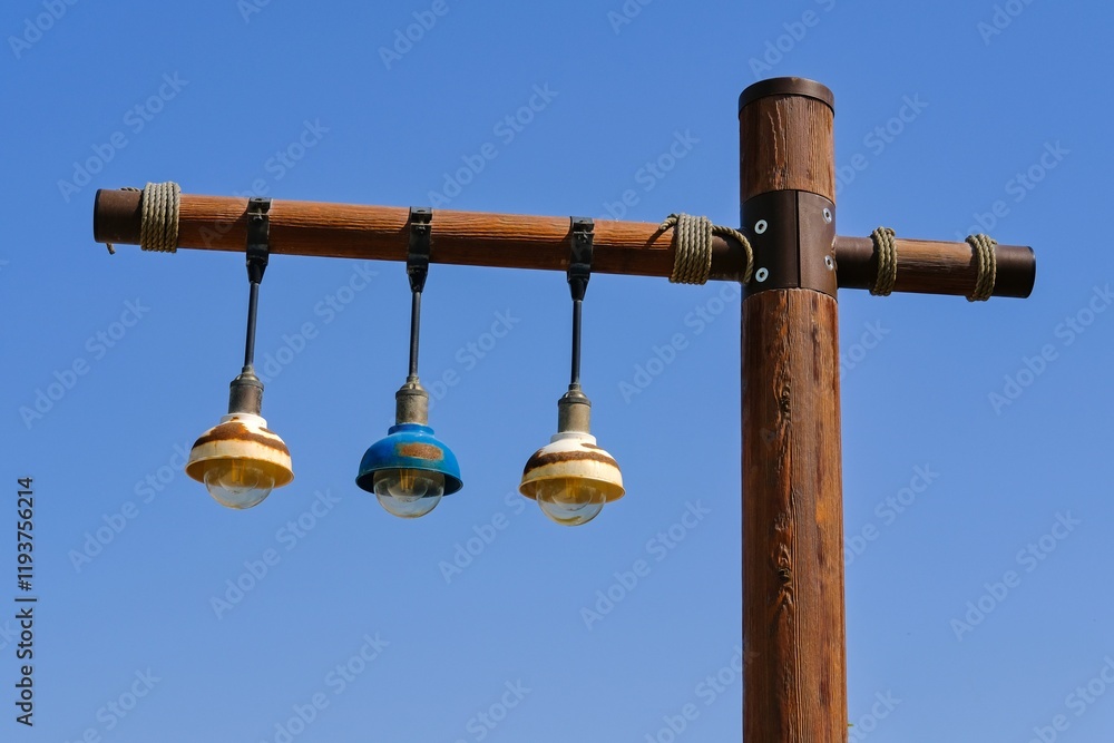 Fototapeta premium Wooden street lamppost with hanging three vintage weathered rusty lamp bulb lantern in trendy eco style on background of blue sky during day