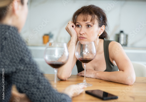 Relaxed middle-aged woman sitting at table with a glass of wine while having conversation with a friend at home photo