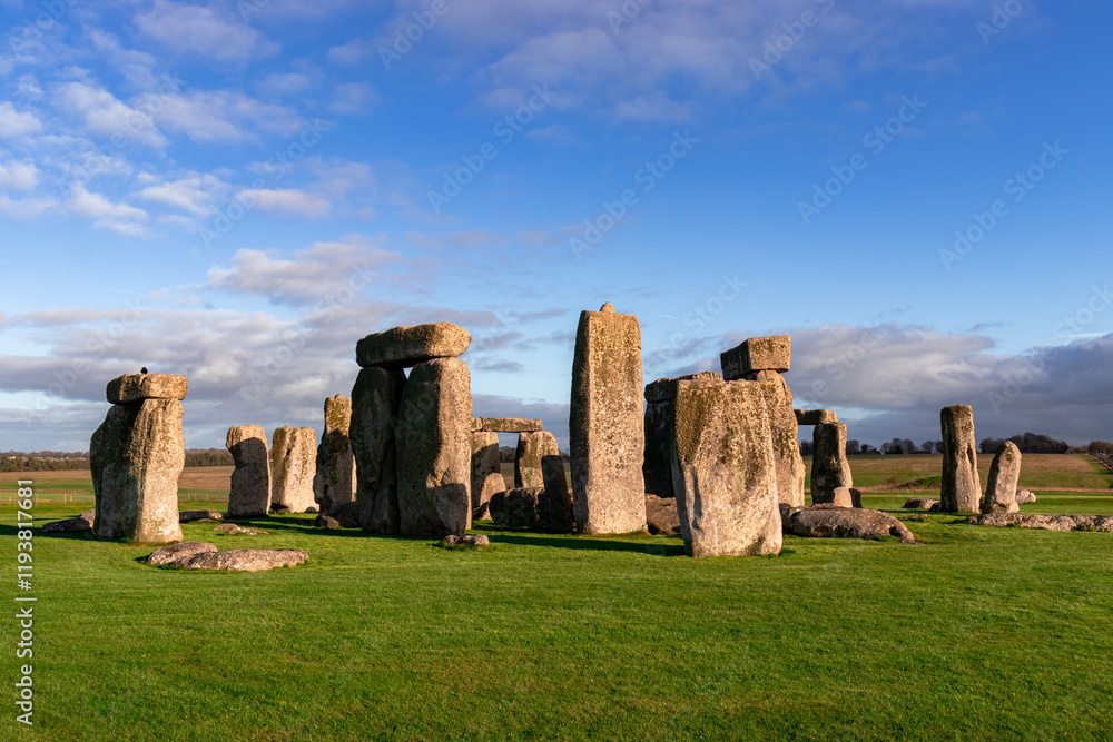 Naklejka premium A captivating view of Stonehenge, the iconic monument in England, illuminated by warm sunlight against a vibrant sky. The ancient stones stand tall on a lush green field
