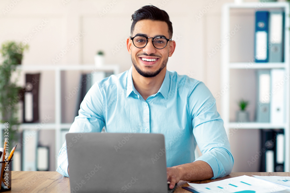 Happy young Arab businessman in glasses sitting at desk, working on laptop at company office. Successful smiling CEO having online job, typing on pc keyboard, having video call Happy young Arab businessman in glasses sitting at desk, working on laptop at company office. Successful smiling CEO having online job, typing on pc keyboard, having video call
