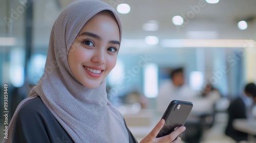 Smiling woman in hijab uses smartphone in a bright, modern office space photo