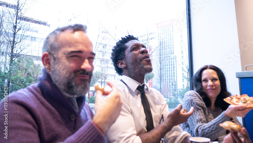 Three colleagues, including a bearded man, laugh and enjoy pizza together, highlighting teamwork, friendship, and a relaxed workplace environment