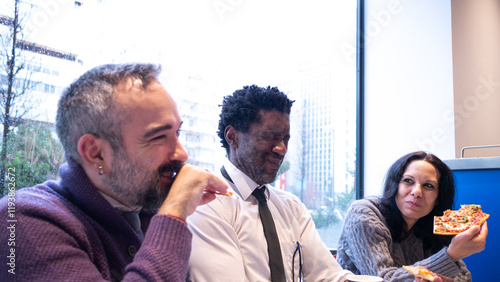 Three colleagues, including a bearded man, enjoying pizza and laughing together in a relaxed setting, capturing teamwork, joy, and a friendly office culture