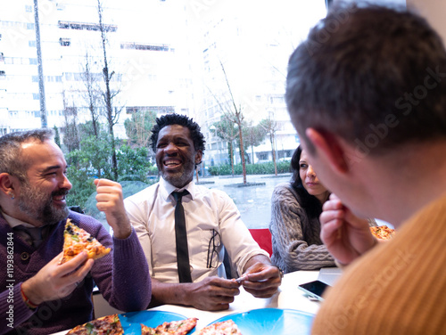 Seen from the perspective of a fourth person in a beige sweater, three colleagues enjoy pizza and laugh together, with a phone on the table. The woman appears slightly more serious
