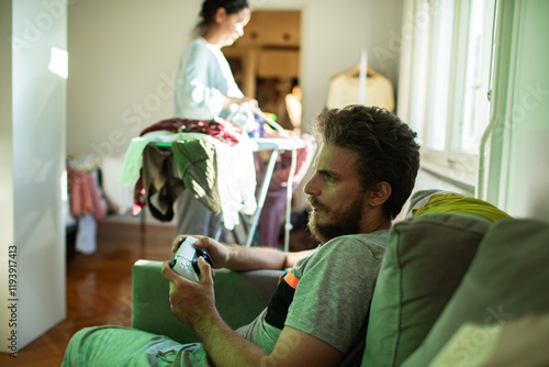 Man playing video games while woman does laundry in background photo