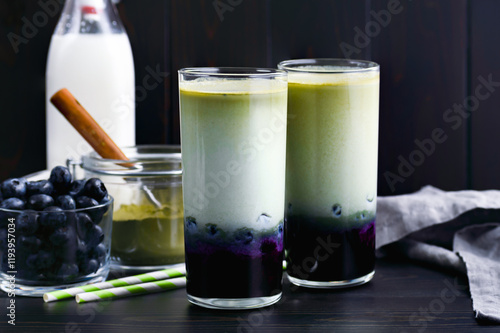Iced Blueberry Matcha Lattes in Tall Glasses: Cold lattes made of layered green tea, milk, and blueberries with paper straws and ingredients nearby