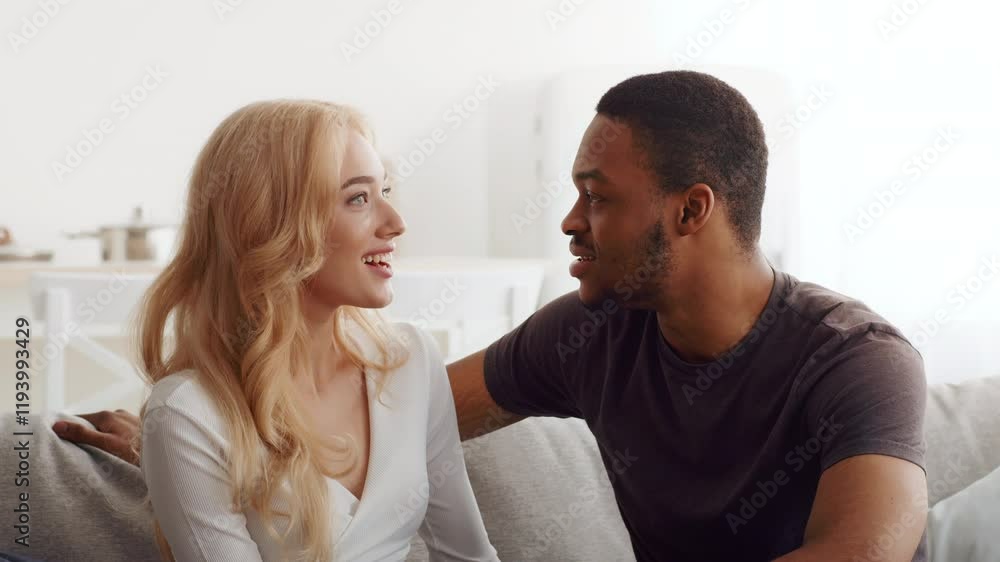 A biracial couple sits closely together on a couch, exchanging smiles and warmth. They enjoy a serene moment, surrounded by the comfort of their home, embodying love and connection. A biracial couple sits closely together on a couch, exchanging smiles and warmth. They enjoy a serene moment, surrounded by the comfort of their home, embodying love and connection.