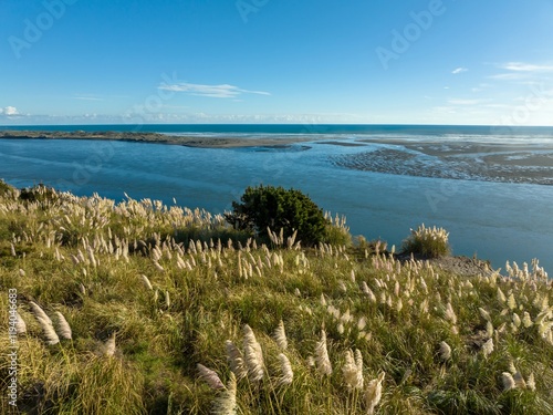 Panoramic view of a New Zealand river meeting the ocean. Fluffy grasses on a hillside overlook the tranquil scene. Beautiful natural landscape. Waikato River, Port Waikato, Waikato, New Zealand photo