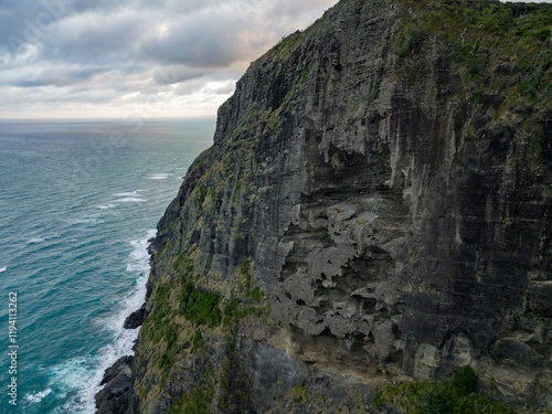Dramatic coastal cliff face, sculpted by the ocean. Waves crash against the rocky shore. Nature's powerful artistry. Mercer Bay Loop, Piha, Auckland, New Zealand