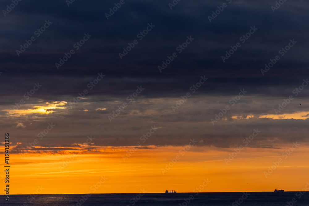Obraz premium Stunning sunset over ocean, dramatic clouds, silhouetted ships on horizon, creating serene, majestic scene