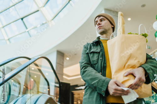 Responsible consumer holding paper bag with fresh groceries on escalator, monitoring rising prices, adjusting budget, and maintaining balanced diet. photo