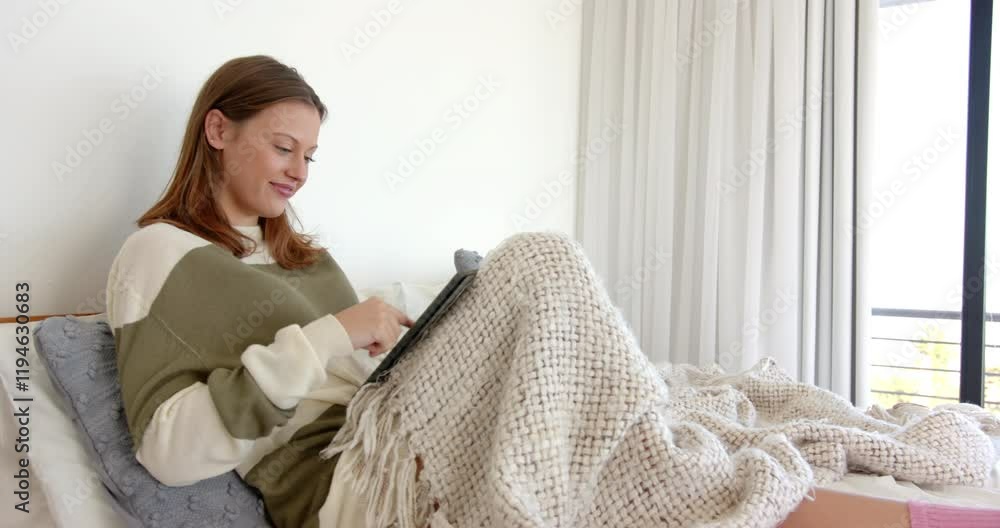 Relaxing on bed, young woman using tablet and smiling under cozy blanket Relaxing on bed, young woman using tablet and smiling under cozy blanket
