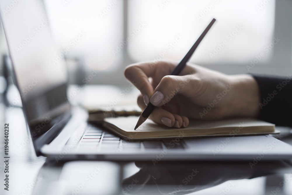 The image captures a woman's hand in close view as it writes in a notepad on a chic laptop, background out of focus The image captures a woman's hand in close view as it writes in a notepad on a chic laptop, background out of focus