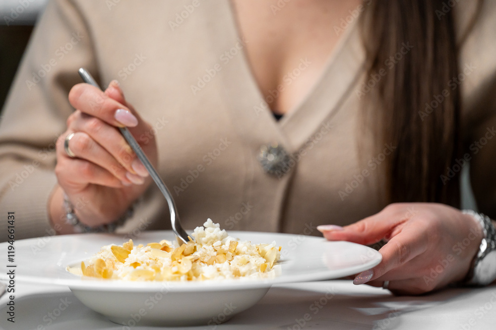 A close-up of a woman's hand holding a fork, serving a dish with rice and nuts on a white plate. The focus is on the meal and the elegant presentation, highlighting dining experience. A close-up of a woman's hand holding a fork, serving a dish with rice and nuts on a white plate. The focus is on the meal and the elegant presentation, highlighting dining experience.