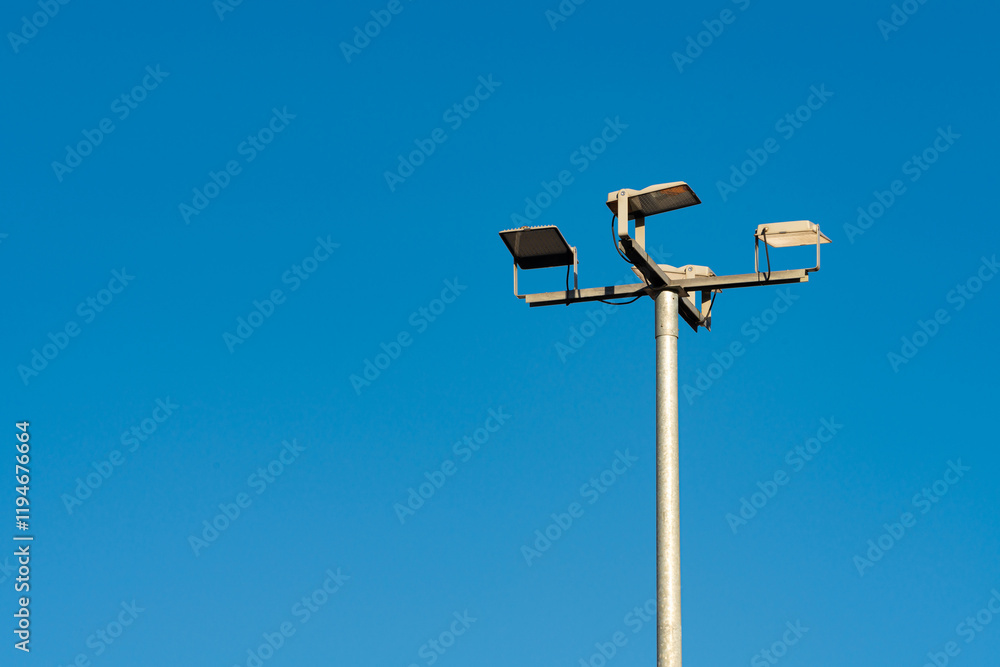 A tall pole supports multiple streetlight fixtures against a bright blue sky. The cloudless backdrop highlights the fixtures, emphasizing urban infrastructure and public safety measures. A tall pole supports multiple streetlight fixtures against a bright blue sky. The cloudless backdrop highlights the fixtures, emphasizing urban infrastructure and public safety measures.