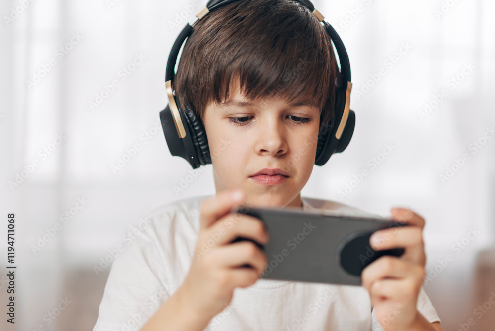 Cute boy with headphones playing games on his phone at home. Happy child looking at his smartphone. Cute boy with headphones playing games on his phone at home. Happy child looking at his smartphone.