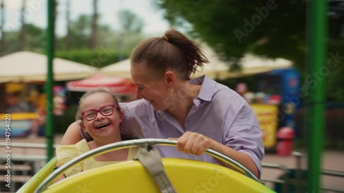 Daughter with Down syndrome in yellow dress and mom in purple shirt laugh together while seated on amusement ride. Scene highlights joy, bonding, and shared adventure