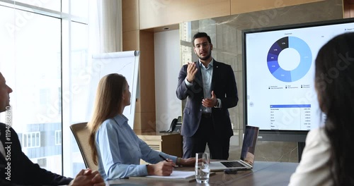 Arabian presenter standing near digital display with charts and data, addresses small group of colleagues, negotiating, discussing financial or business analytics, gathered in modern conference room