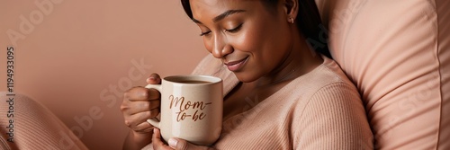 Smiling pregnant woman in a cozy outfit holding a mug labeled Mom to be gently cradling her belly, seated on a couch. Perfect for maternity and lifestyle