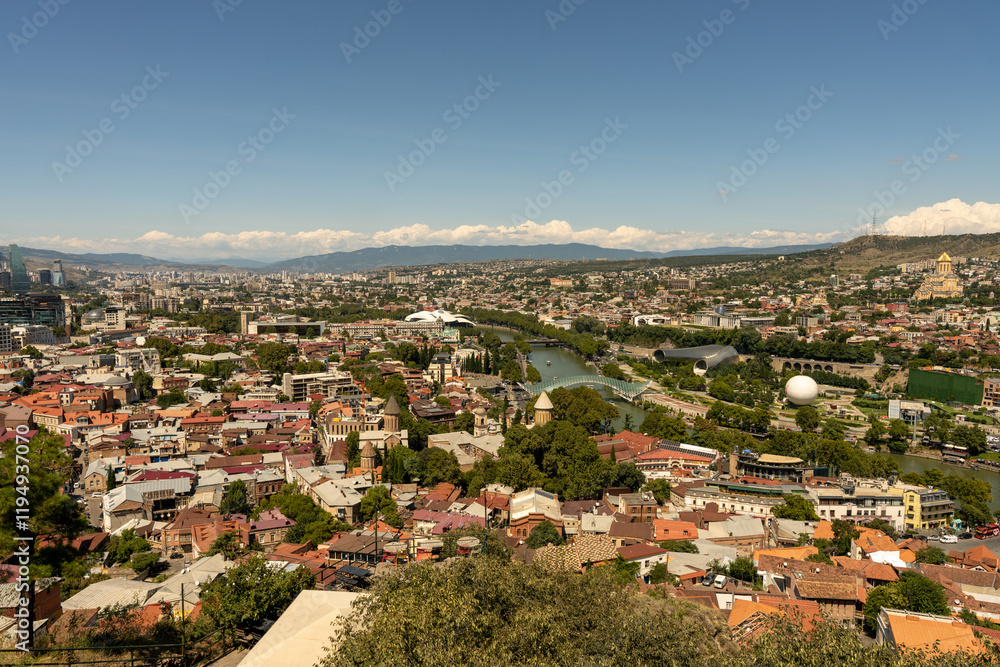 A city view with a river running through it. The city is full of buildings and trees. The sky is clear and blue
