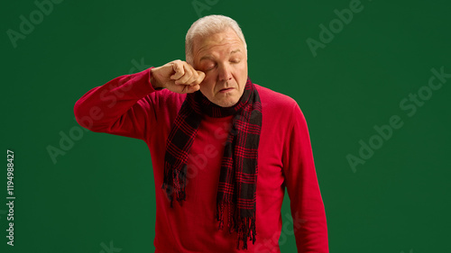 Senior man in red sweater and scarf swiping tears, suffering from seasonal allergy symptoms on green studio background. Concept of illness, medicine, treatment, health care photo