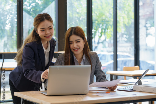 Two Asian businesswomen sit at desks discuss project details, share opinions, and work on collaborative tasks.