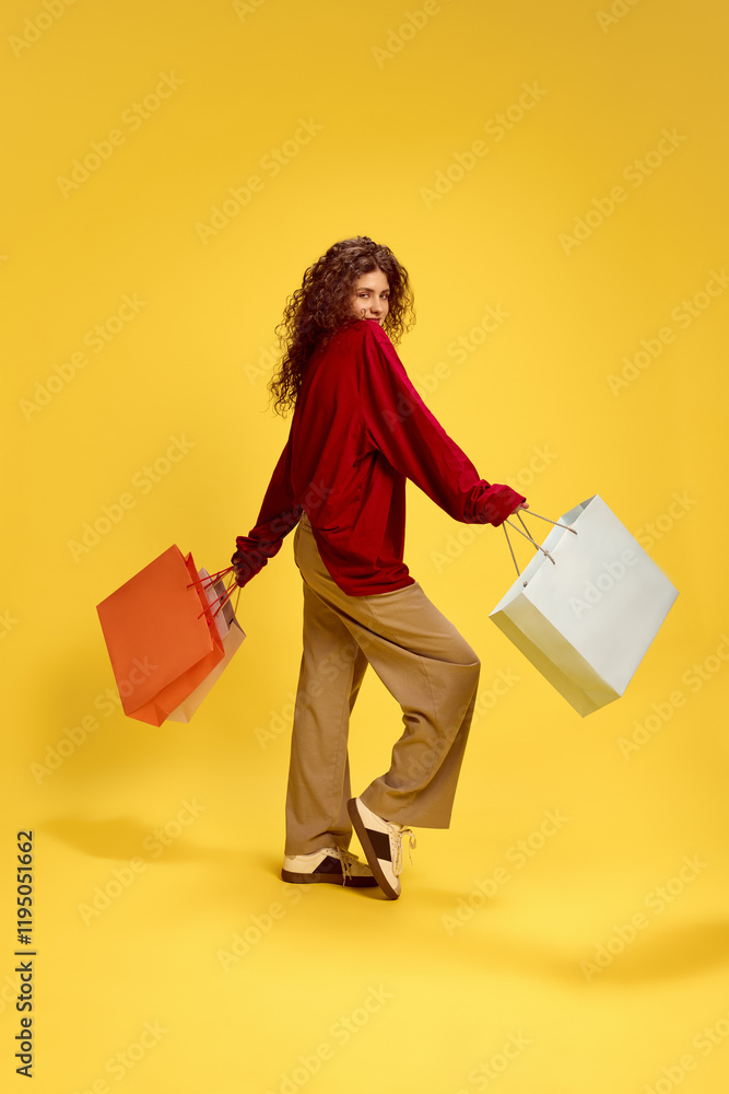 Rear view of woman, looking at camera throw shoulder with playful expression holding shopping bags against yellow studio background. Concept of human emotions, self-expression, fashion and style. Rear view of woman, looking at camera throw shoulder with playful expression holding shopping bags against yellow studio background. Concept of human emotions, self-expression, fashion and style.