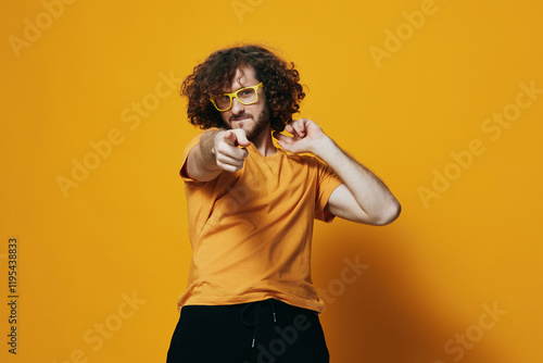 Young man with curly hair in yellow glasses pointing at the camera, wearing an orange t shirt against a vibrant yellow background, evoking a playful and confident mood photo