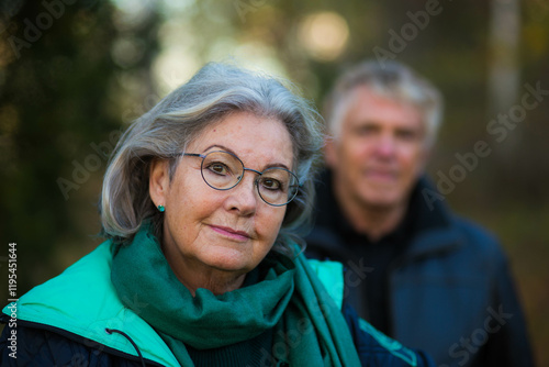 A portrait of an older woman and her husband, out of focus, in the background outside in the Swedish nature