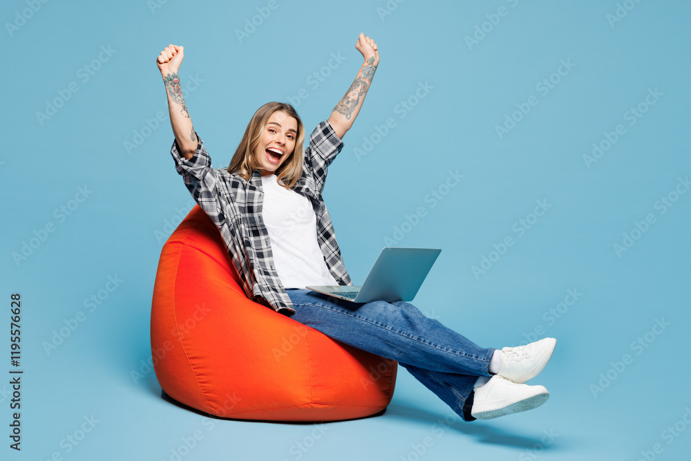 Full body young fun IT woman wears checkered grey shirt casual clothes sit in bag chair hold use work on laptop pc computer do winner gesture isolated on plain blue cyan background. Lifestyle concept. Full body young fun IT woman wears checkered grey shirt casual clothes sit in bag chair hold use work on laptop pc computer do winner gesture isolated on plain blue cyan background. Lifestyle concept.