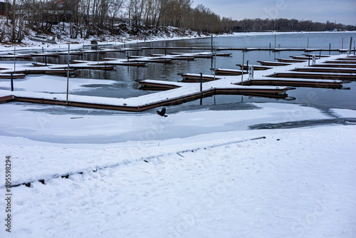 Frozen water, river with landing slip, wooden dock, floating pontoon for boats. The water is partially covered in ice and snow. A lone bird flies over the frozen surface.  photo