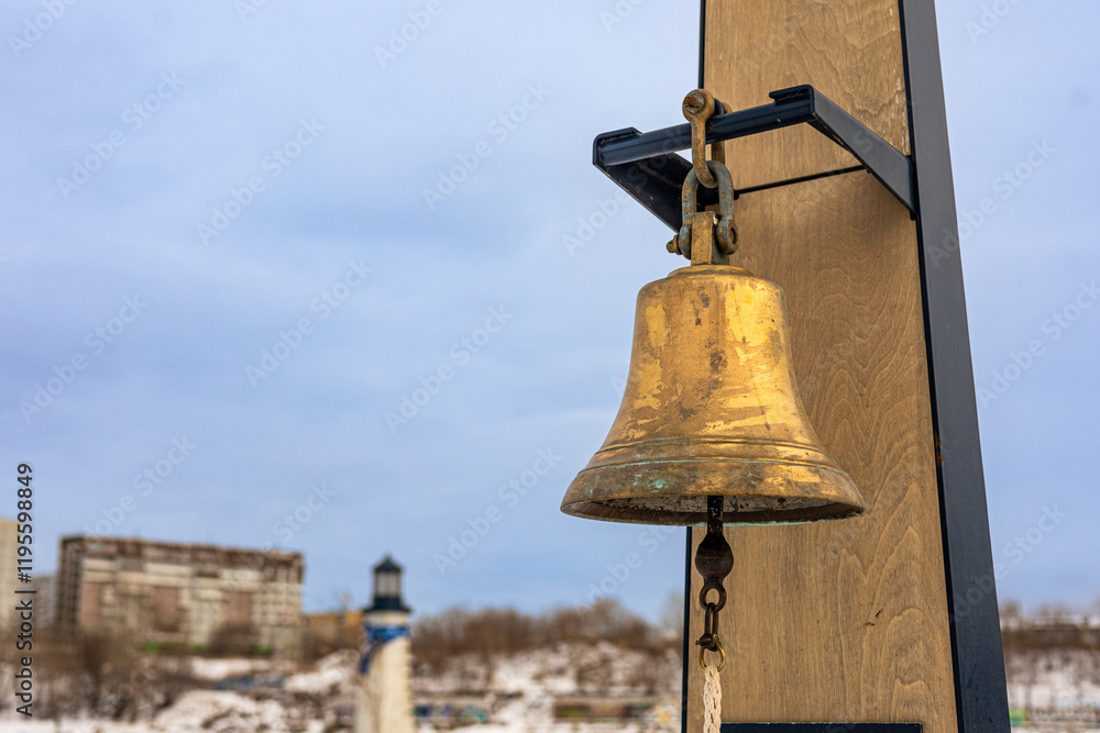 Obraz premium Brass bell hanging from a wooden frame. Ships bell, rynda. Defocused snowy landscape with lighthouse and buildings on cold full winter day.