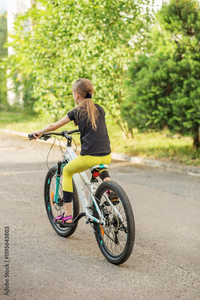 Fototapeta premium Preteen girl riding bicycle along tree-lined path during sunny weather