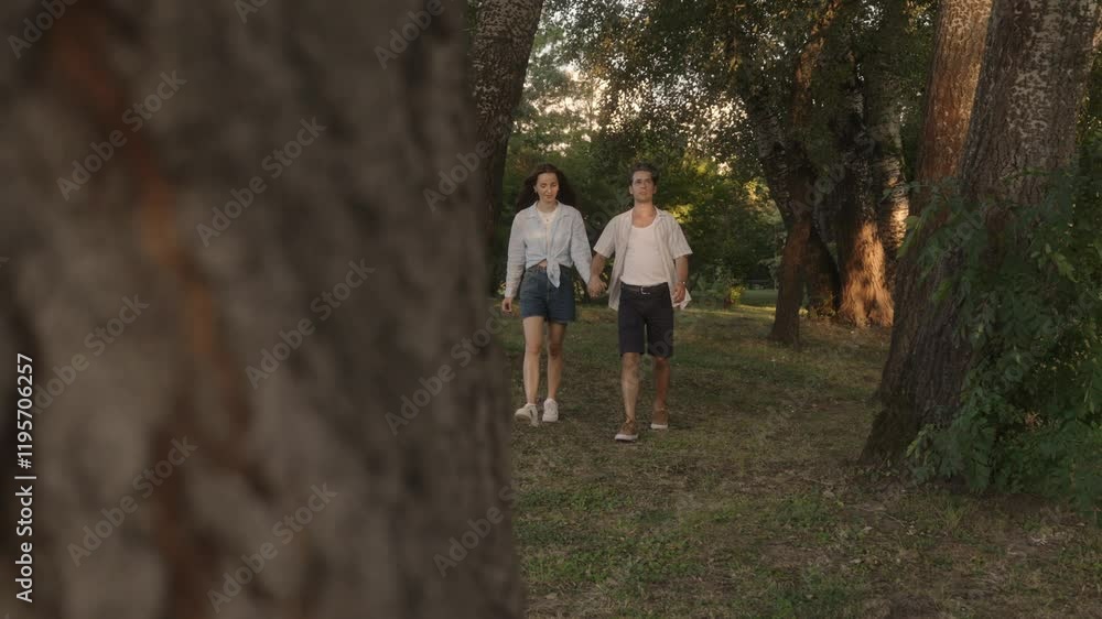 A young couple holding hands and walking through a park between the trees. Romantic summer evening with soft golden sunlight. A young couple holding hands and walking through a park between the trees. Romantic summer evening with soft golden sunlight.