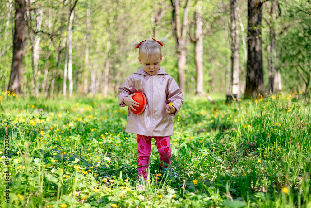 A little girl of 2-3 years old stays in the forest and enjoys nature. Background of a green forest. The girl holds tightly in her hand an orange jar in which she collects the flowers she likes.