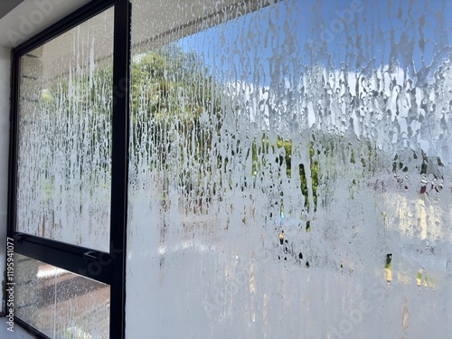 An aluminum-framed residential window as seen from inside the home, covered in a layer of suds during a professional pressure pre-treatment wash.